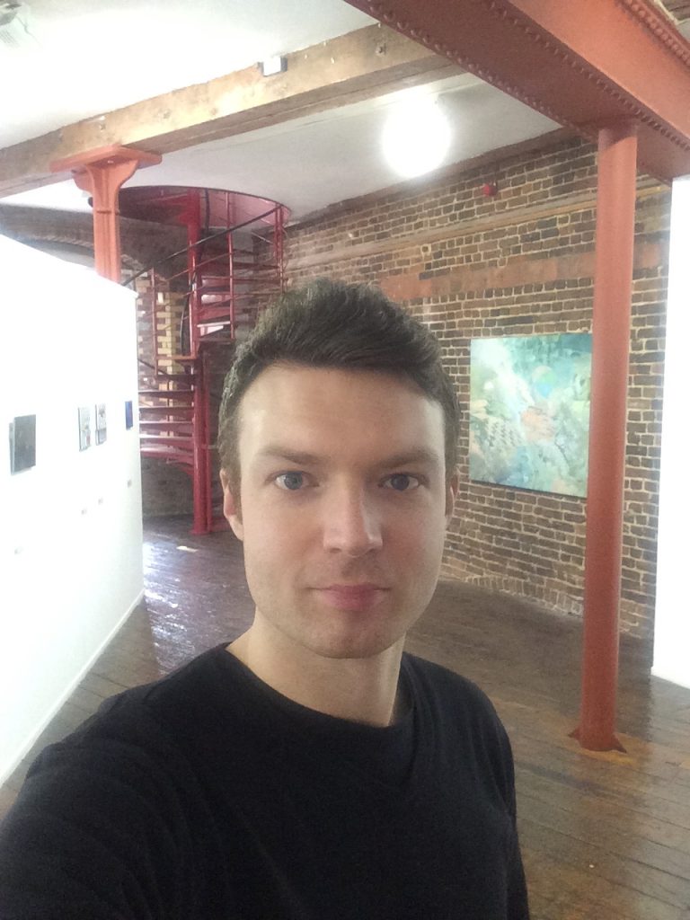 Photo of a white male with short brown hair. He is wearing a black long sleeved jumper. Stood inside an exposed brickwork room.