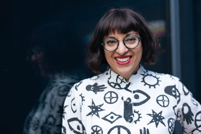 Head and shoulders shot of Binita Walia. She is smiling and is wearing round glasses with a white top covered in black line drawn symbols.