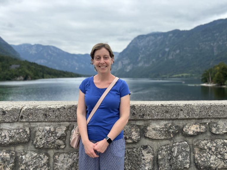 Rachel Lloyd smiling and standing in front of a wall with a picturesque late and mountains in the background.